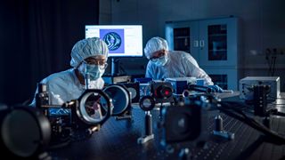 Two people wearing blue clean suits and masks bend over a laser table full of mirrors, lenses, and lasers