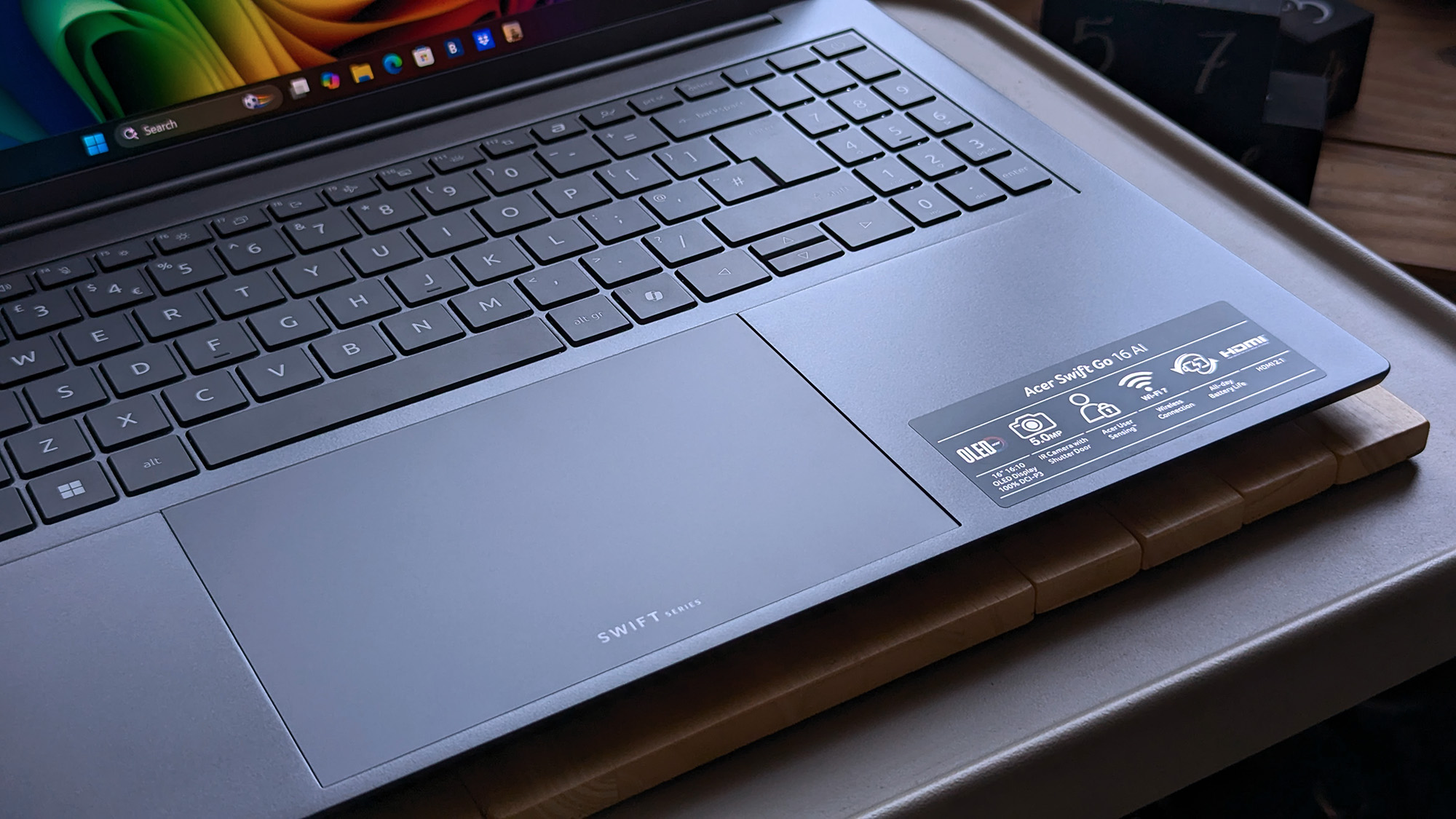 A close-up of the Acer Swift Go 16 AI laptop showing its keyboard, large touchpad, and feature stickers highlighting the OLED display, 5 MP webcam, PurifiedVoice, Wi&amp;#8209;Fi 7, and long battery life.