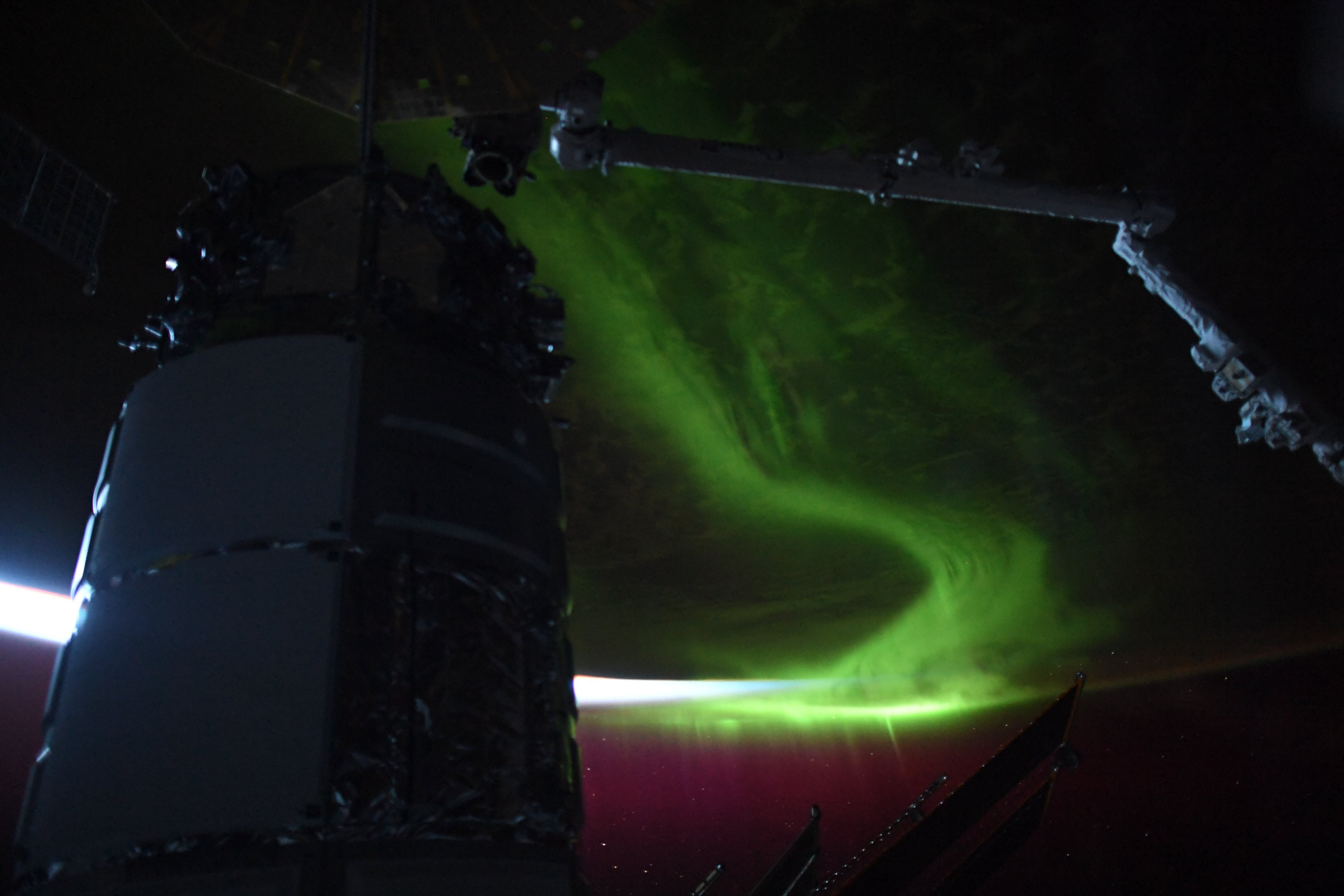the dark, nearly silhouetted shape of a cylindrical spacecraft rises from the left, briefly reflecting the slice of white light curved from the horizon of Earth from space, seen above. a robotic arm reaches to the mass from the right, as behind a swath of green aurora smears across the dark face of the planet.