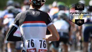 A rear view of a white male road cyclist, wearing a black, white, red and blue jersey with race numbers on the pockets, and one black arm warmer on his left arm, while his right arm is bare