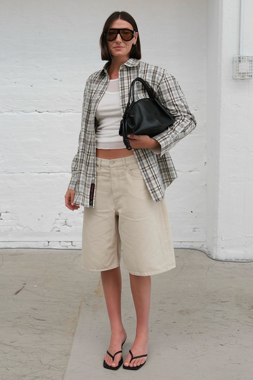 A woman wearing a plaid shirt, white tank top, tan shorts, and black thong sandals