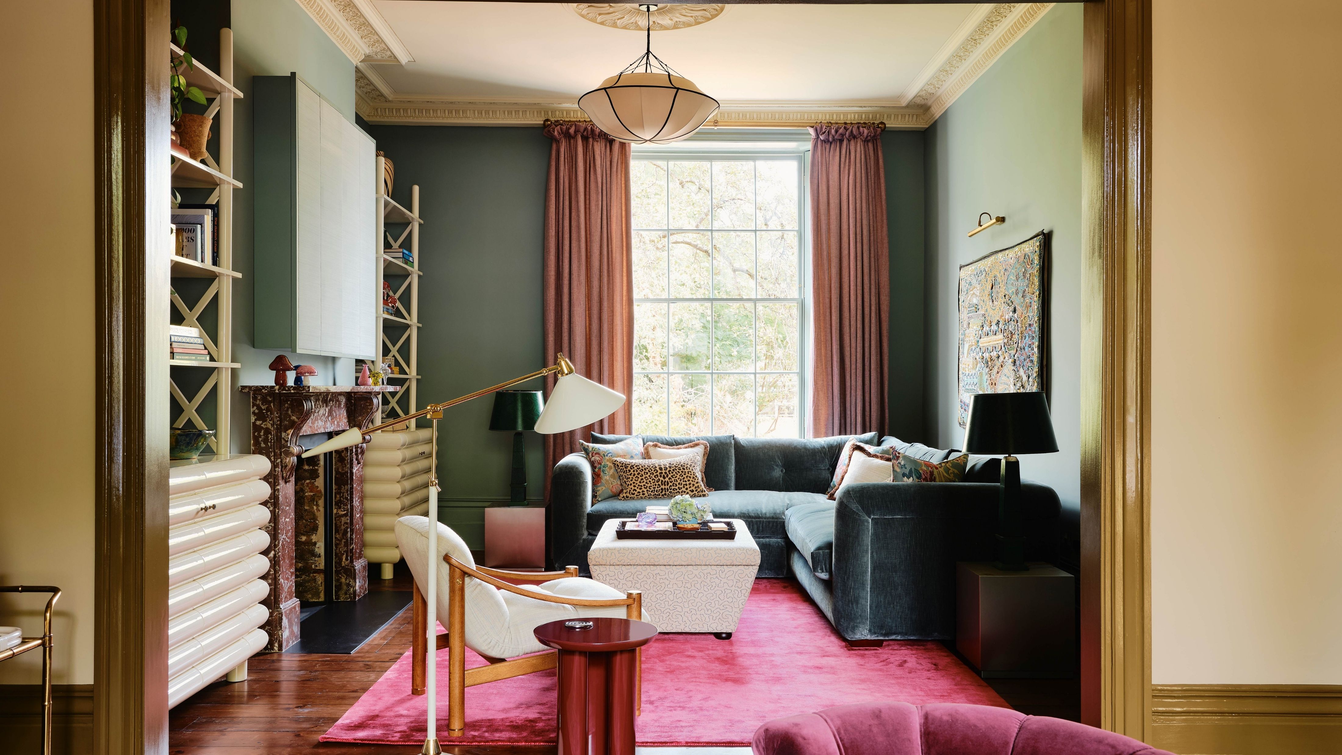Weathered teal velvet sofa pictured in a colorful pink and burgundy accented living room with wooden floors and a pendant light hanging overhead