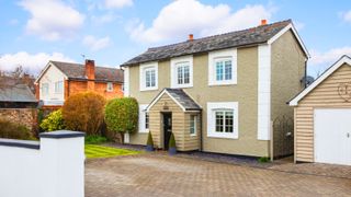 wooden clad porch on stone coloured rendered property with tiled roof, white windows and reveals, garage with wooden door, paved driveway and small front lawn