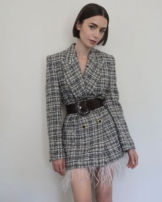 Lily Collins stands in front of a plain wall wearing a belted blazer dress with a feather hem