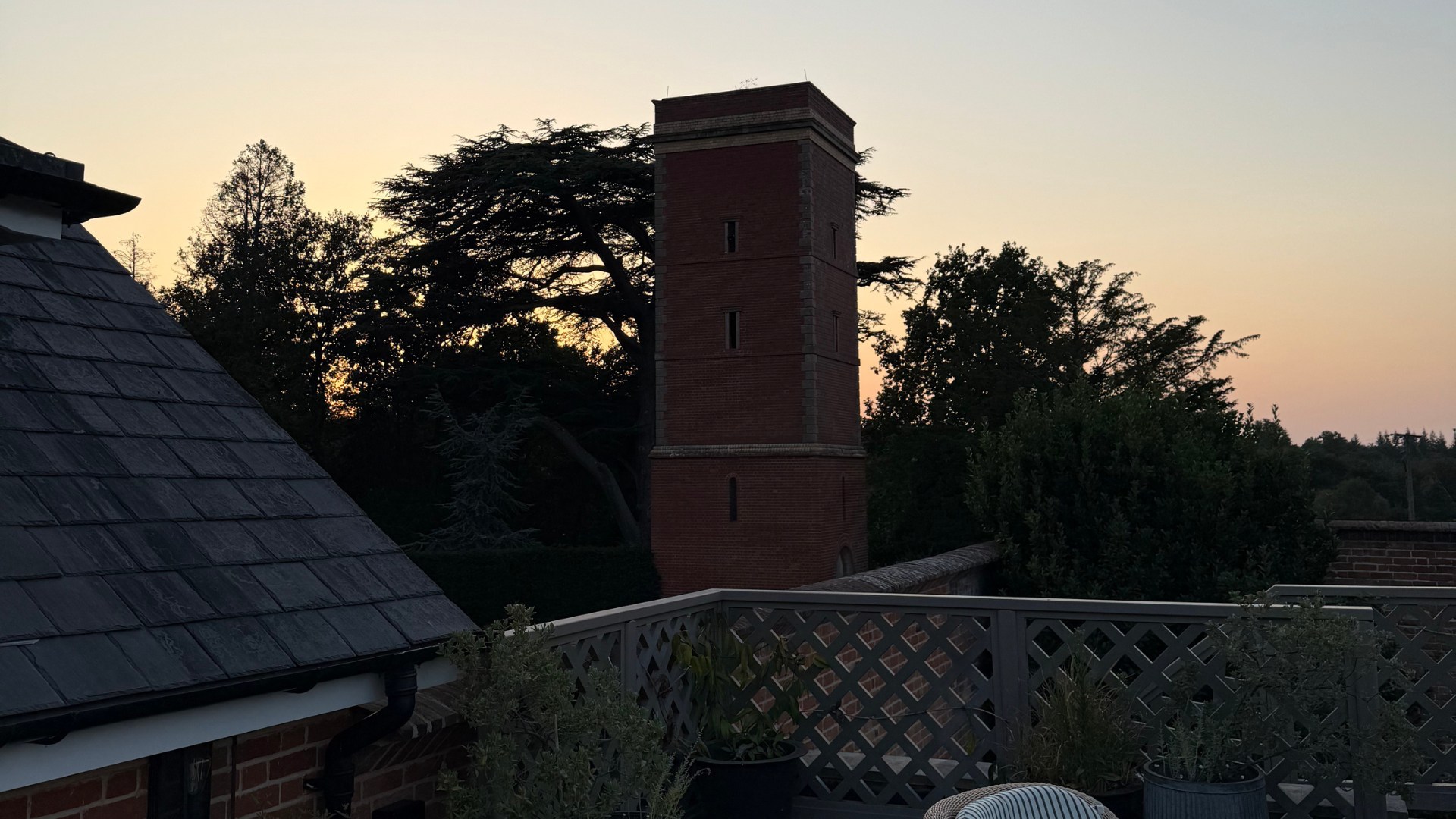 A twilight view from the cottage terrace, looking towards a tall red-brick tower framed by trees and silhouetted against the evening sky. Slate rooftops and potted plants are visible in the foreground.