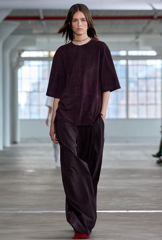 Model walking the Tibi spring runway show wearing a suede eggplant top with matching trousers and red ballet flats.