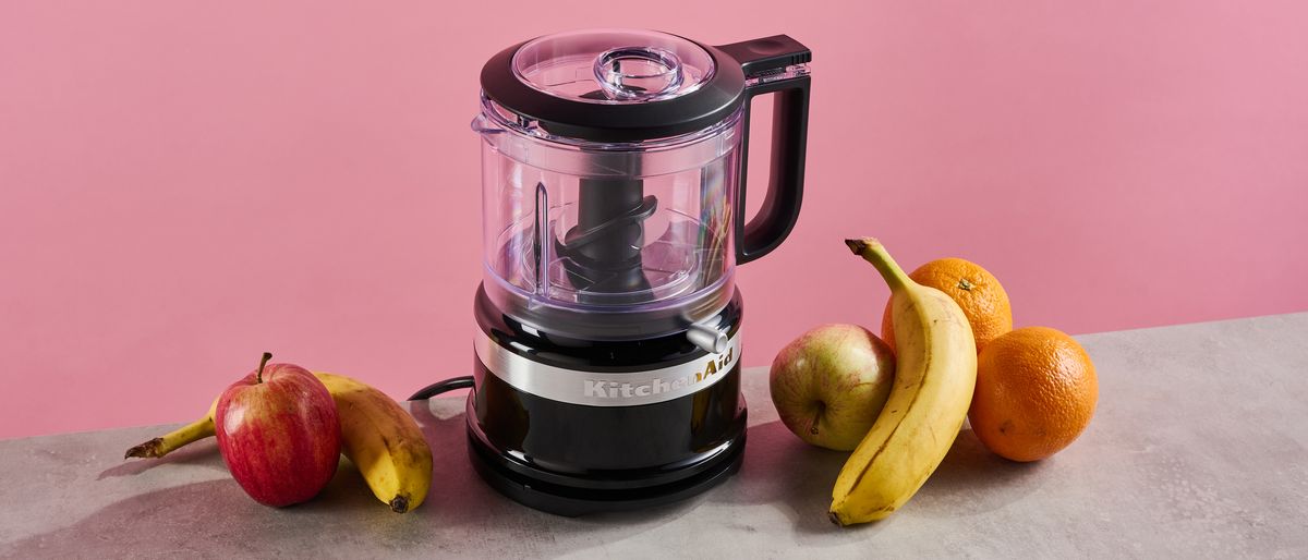 A black gloss KitchenAid 3.5 cup mini food chopper sits on a grey stone-effect surface. The background is pink, and there are apples, bananas and pears on the surface around the chopper.