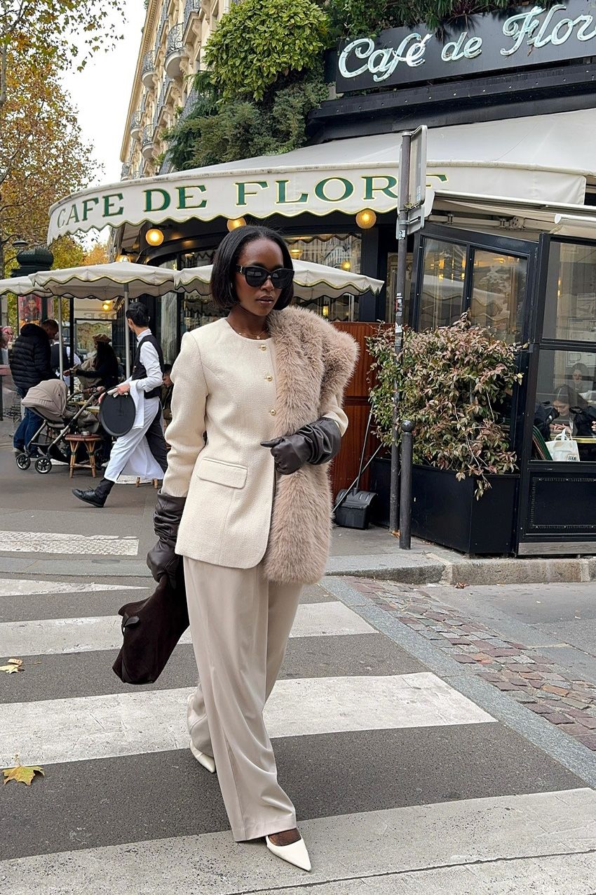 A woman crossing the sidewalk in Paris wearing black sunglasses with a white blazer, brown fur scarf, brown leather gloves, white wide-leg trousers, white pointed heels, and a brown handbag.