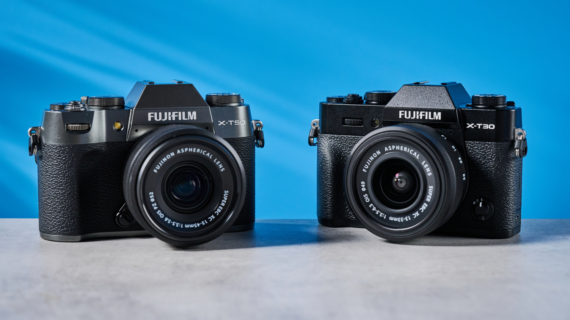 The Fujifilm X-T30 III next to the X-T50 on a stone surface with a blue background