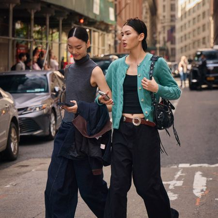 Two Women wear black pants and a basic top and one has a sweater on. 