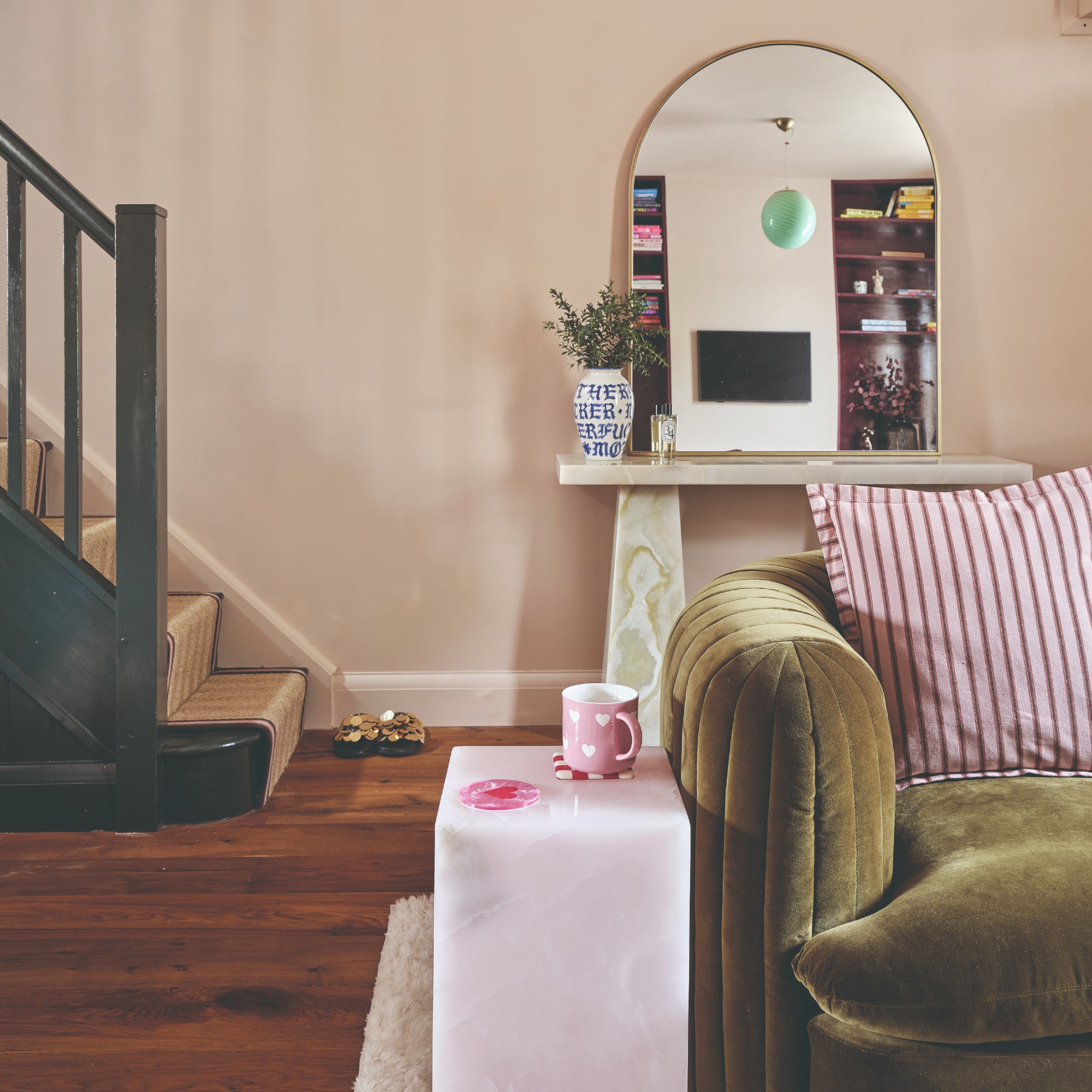 A pink-painted open-plan living room with a green velvet sofa and the TV reflected in the arched mirror placed on a console table behind it