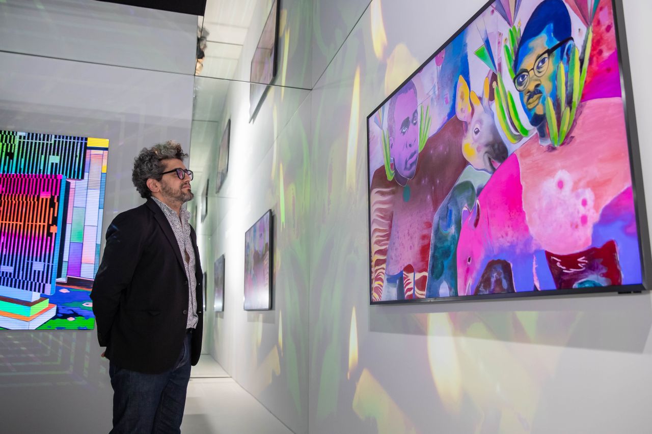 A man dressed in a black tailored jacket, dark denim jeans, and a shirt, wearing black glasses, stands before a television showing a colorful painting depicting a series of people and animal in bright hues.