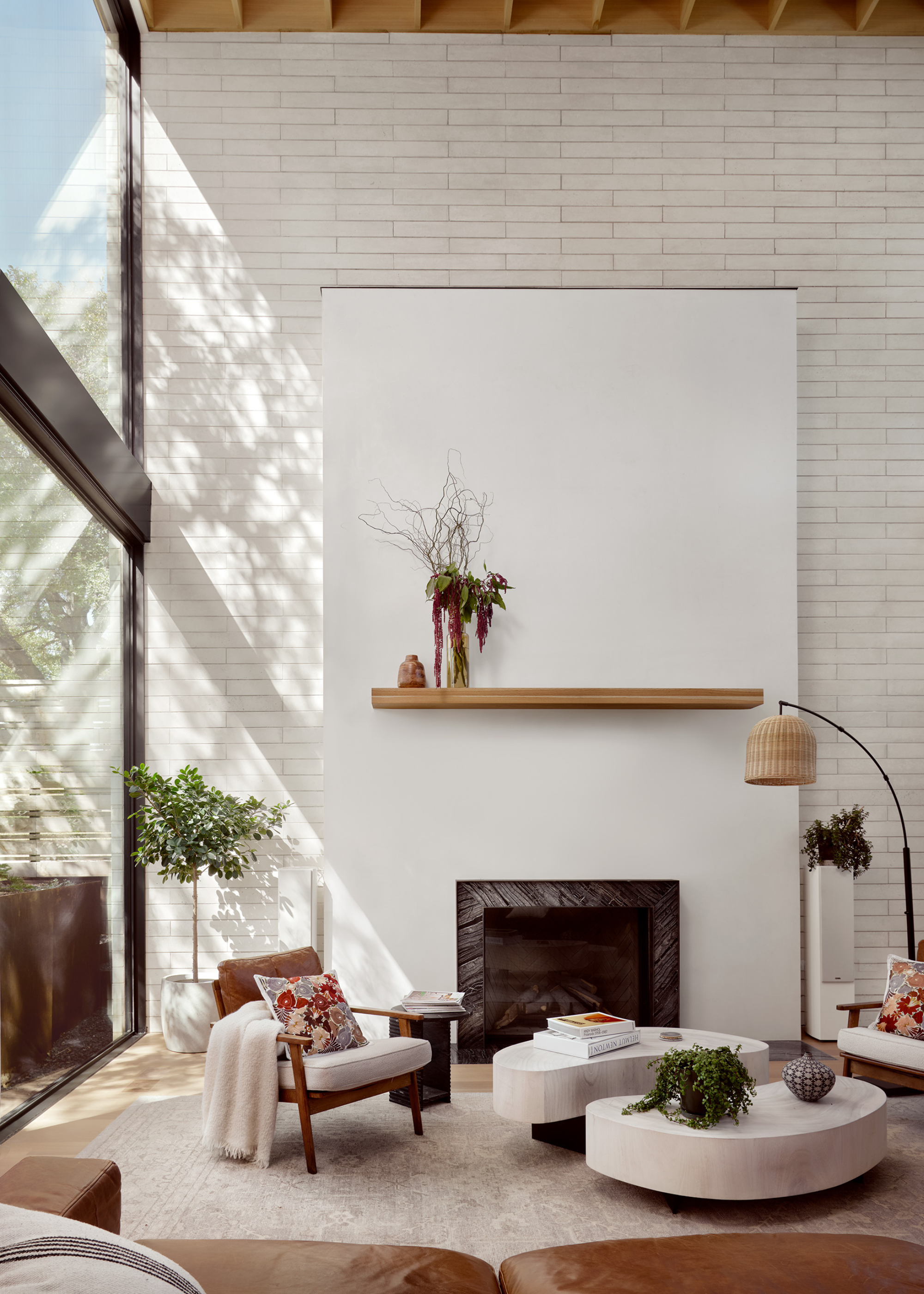 A living room with tall ceilings a white brick back wall, a standing lamp, a potted tree, a set of coffee tables, and a wood chair