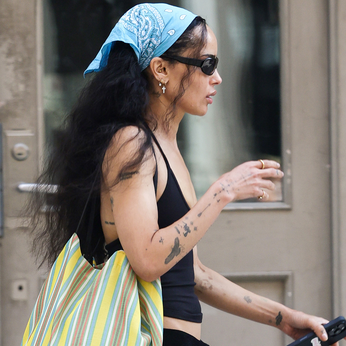 Zoe Kravitz flashes a smile while stepping out in New York City. The American actress carried a large striped tote bag and wore a bandana, black tank top, matching trousers, and sandals.
