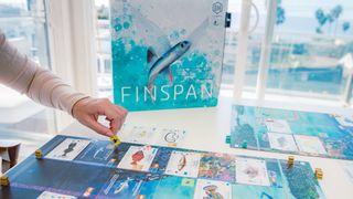 A hand places a Finspan token on the board, with the box in the background