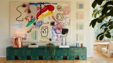 A living room with a white wall holding a large abstract, colorful painting and a set of four square paintings above a green tiled sideboard with a metallic box, a stack of books, a trailing potted plant, a set of candlesticks and a vase