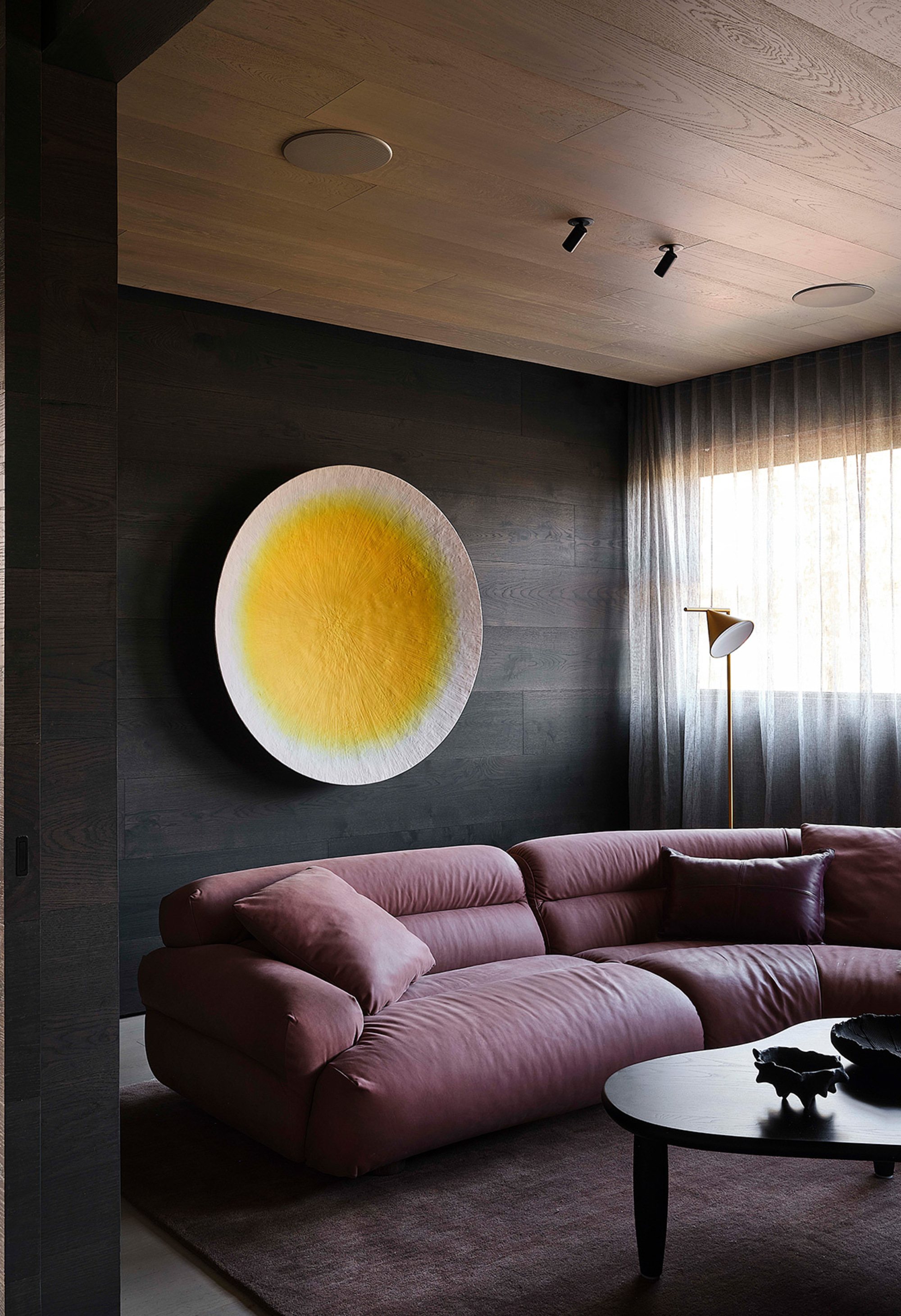 purple sofa in a dark living room with timber ceiling and black charred walls, with dark purple rug, round yellow wall sculpture and black coffee table styled with a vase