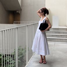 eliza wears a white dress and black flip flops while holding a COS mini studio bowling bag in black