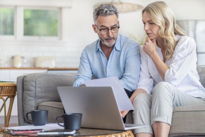 Mature couple using a laptop computer and doing paperwork on the sofa. They are looking worried at a document. The man is wearing glasses