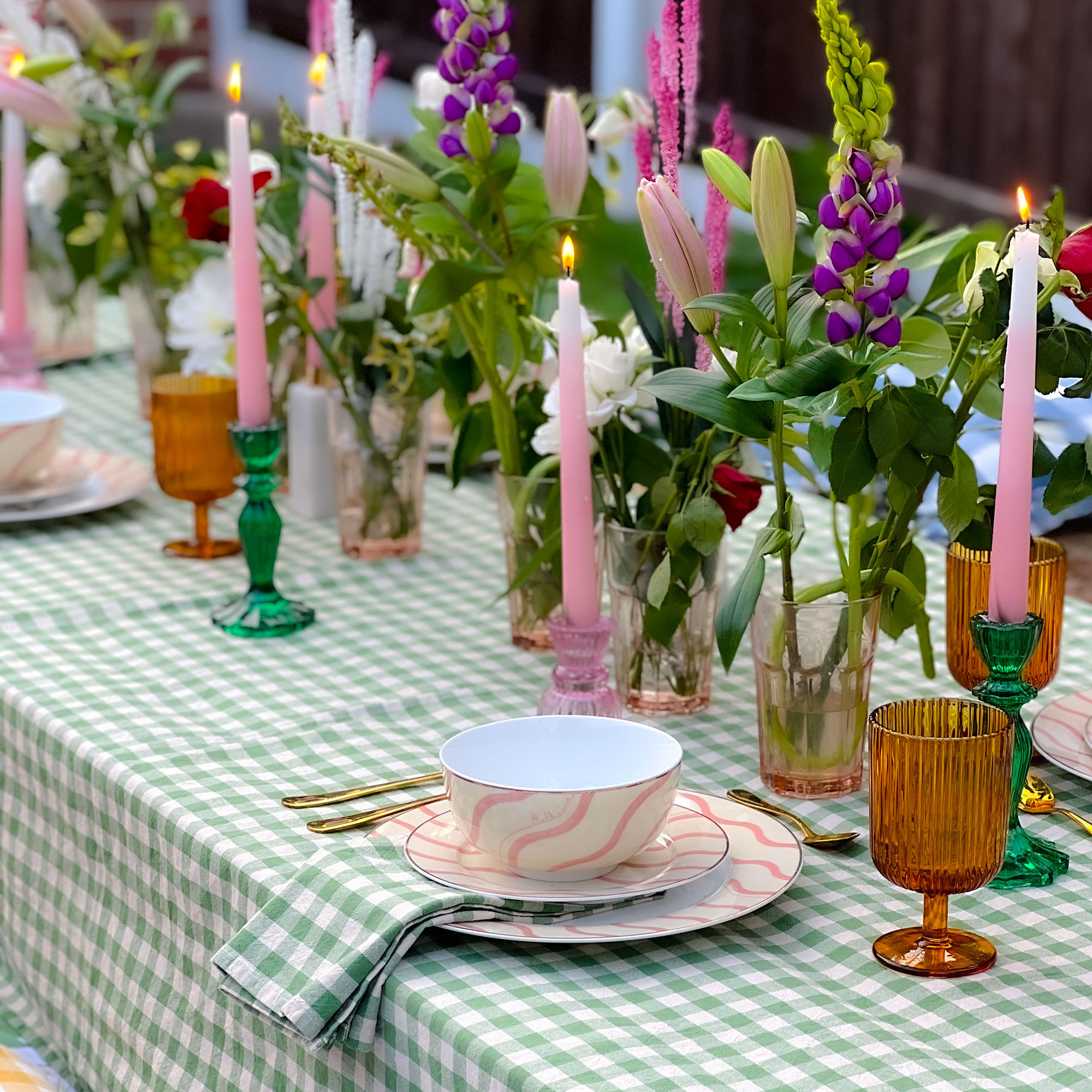 Outdoor table decorated with green and white gingham tablecloth, pink candles, coloured glass candle holders and glasses, mixture of coloured flowers in tumblers