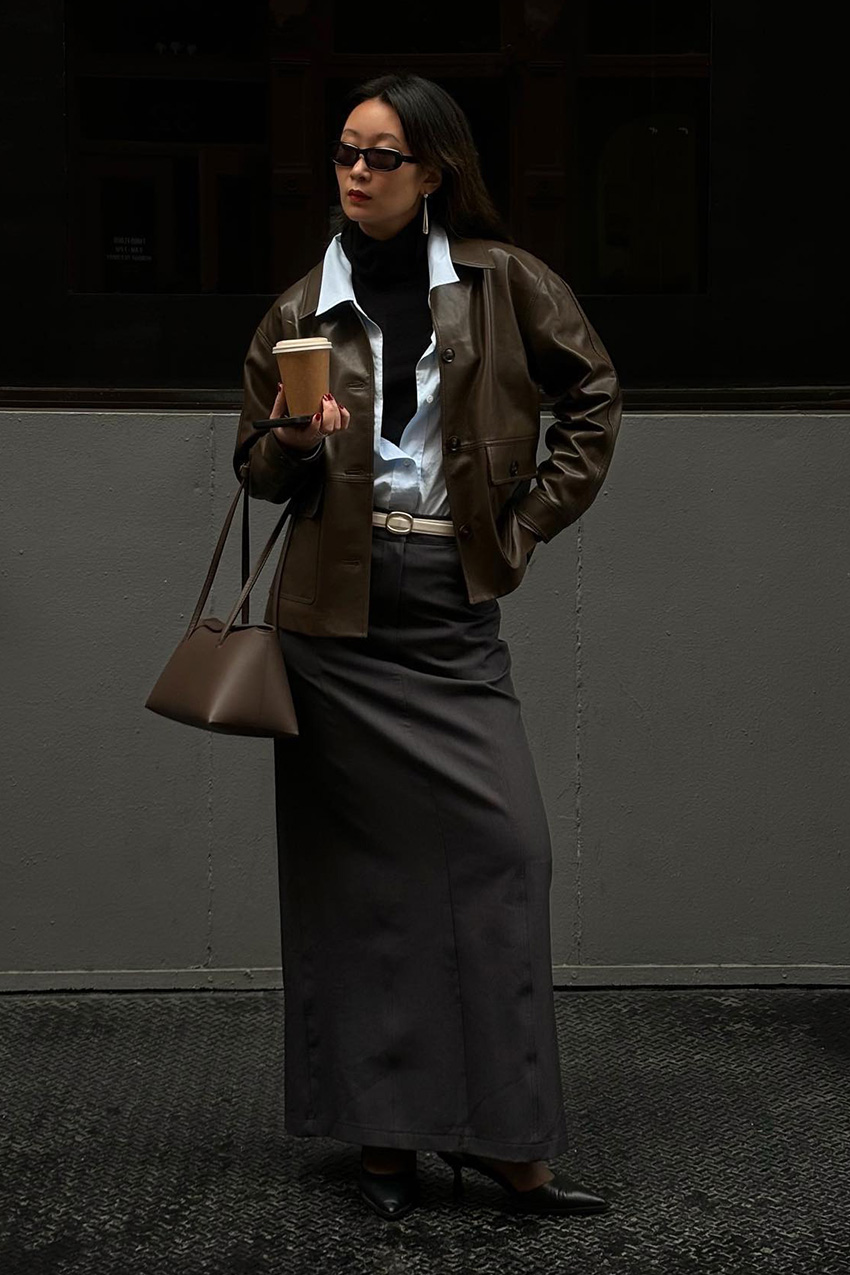 Layering staples for spring 2026 is shown in a street-style photo of a woman standing outside, wearing a brown leather bomber jacket layered over an oversized white button-down shirt, unbuttoned, with a black turtleneck peeking out underneath. The rest of the look includes a gray maxi skirt, a white belt, black pointed heels, a brown shoulder bag, and black sunglasses