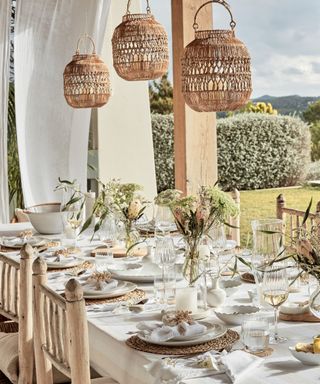 Outdoor dining ideas with three rattan lamp shades above laid out table, view of garden, glassware, white tableware, flowers