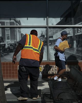 Man wearing orange high-vis near a bench with postal worker passing by