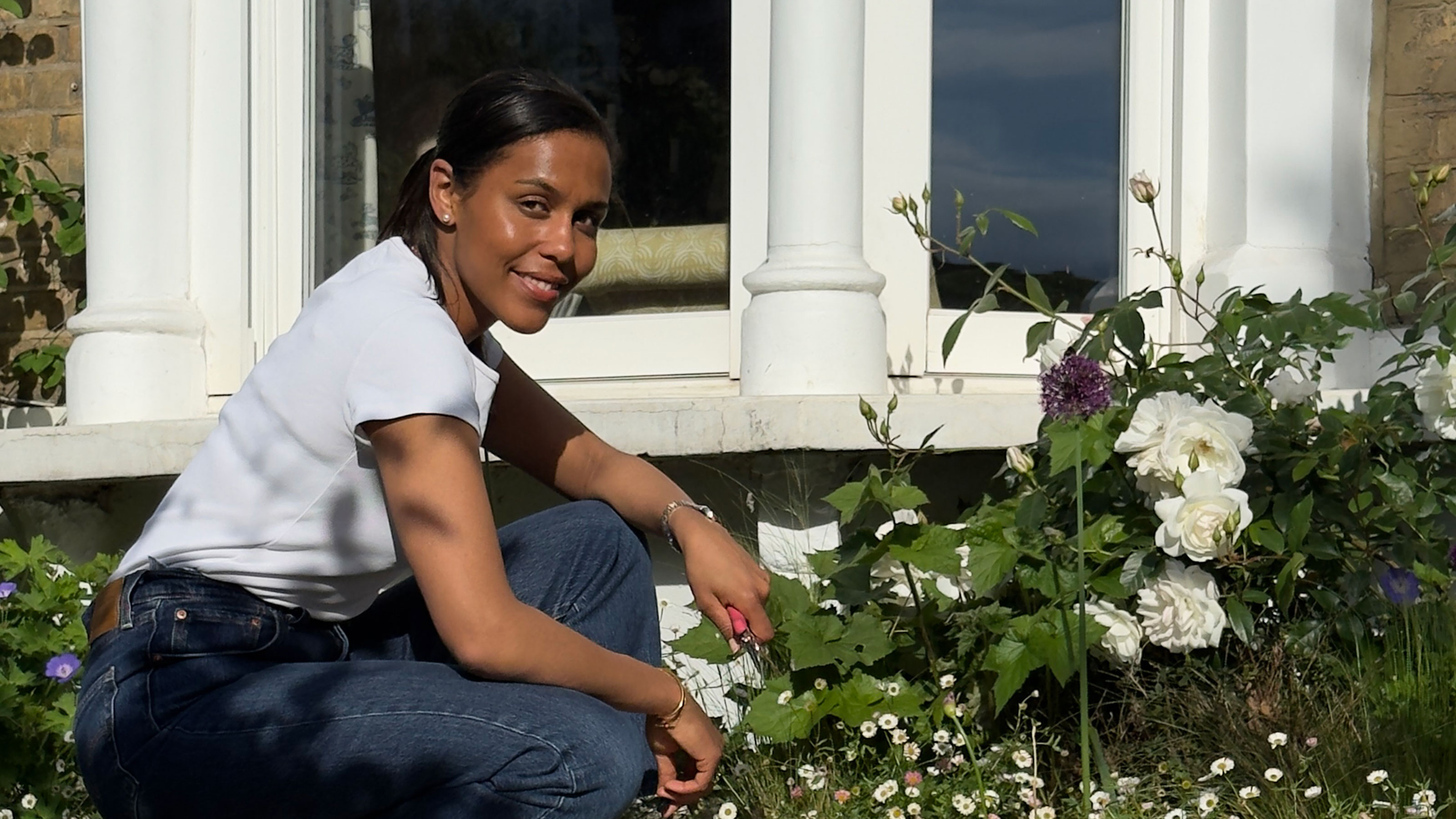 Woman kneeling outside in front of bay window looking at flowering plants