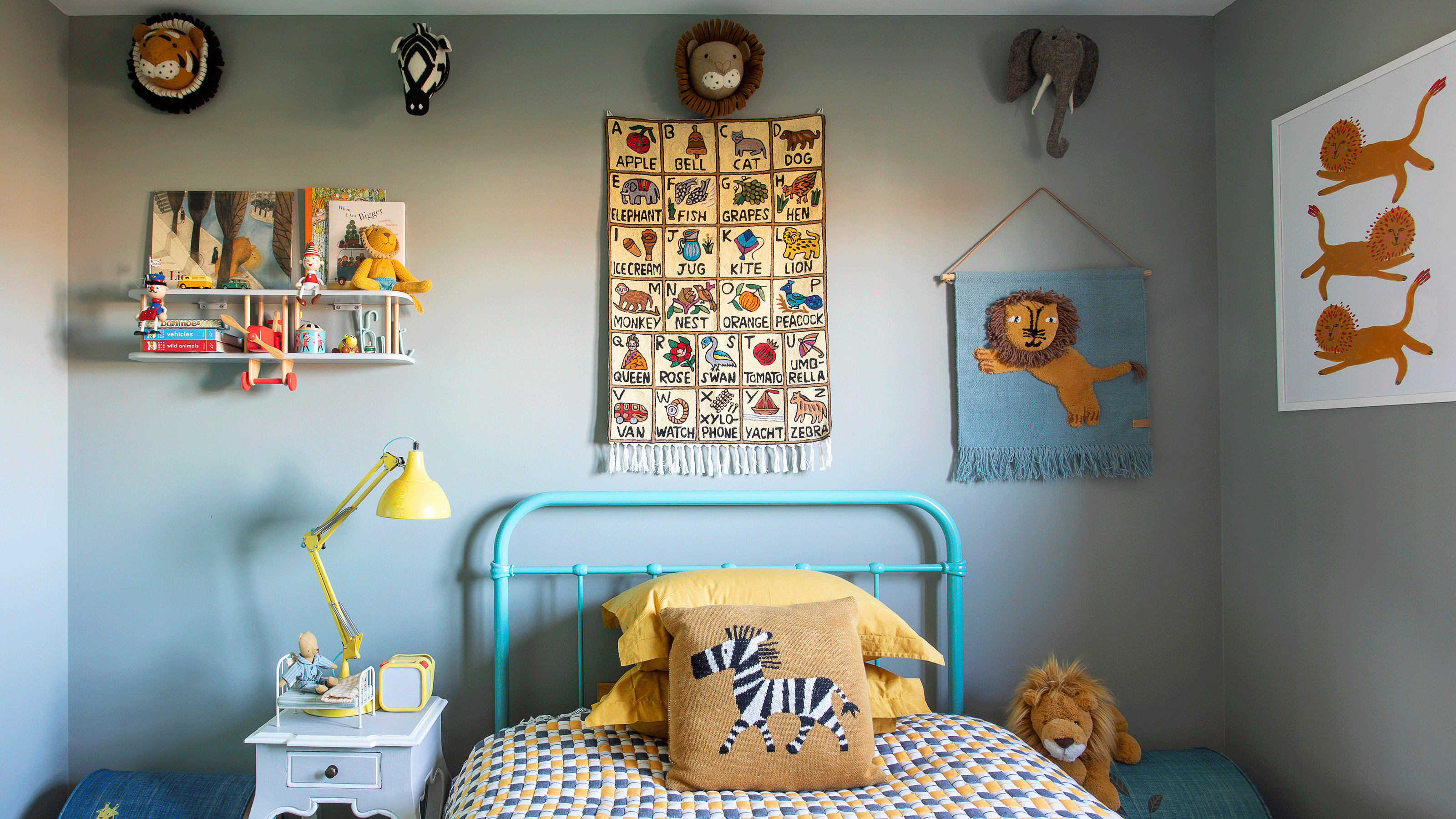 A kid's bedroom painted pale blue with yellow accents, including a bedside lamp, zebra cushion, lion tapestry and alphabet poster