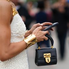 Woman holding home with a black min i handbag in her hand and a gold cuff on her wrist in a white dress
