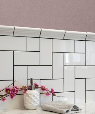 A white subway tiled splashback with dusky pink walls above it and a white surface with a white soap dispenser, towels, and pink blossoms in front of it