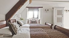 A bright attic bedroom with two twin beds with brown cable knit throws, wooden beds, a white cabinet, white walls, and exposed beams overhead.