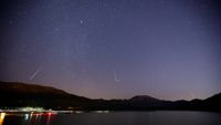 White streaks of meteors are seen against a dark purple night sky with a frozen lake with lights below pushed against silhouetted mountains in the background