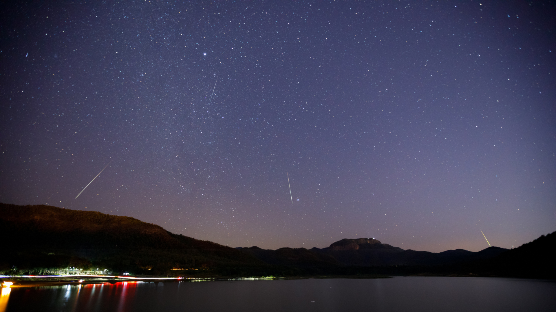 White streaks of meteors are seen against a dark purple night sky with a frozen lake with lights below pushed against silhouetted mountains in the background