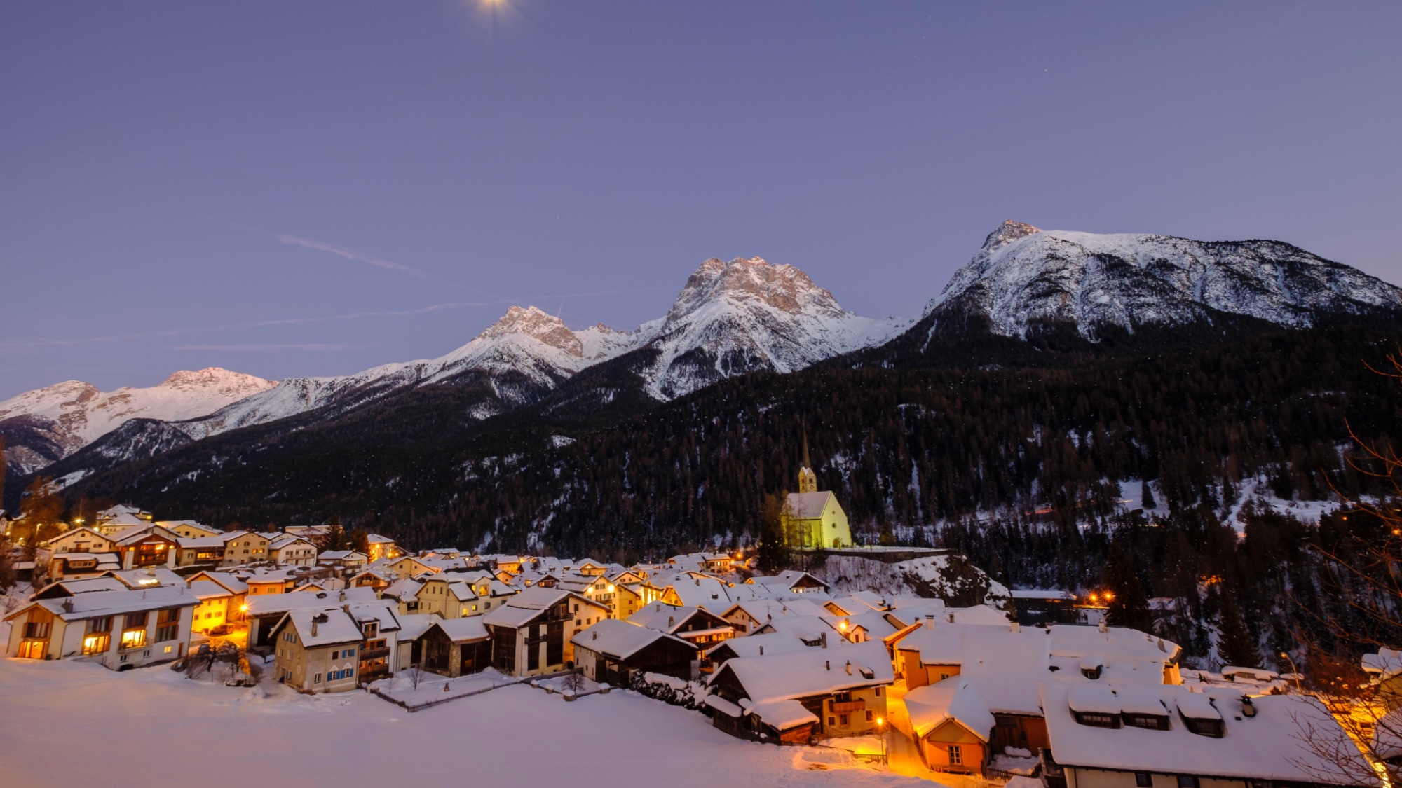 Village of Scuol, Switzerland at dusk, with lights on in houses, and snowy mountains rising behind