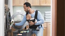 A dad gets clothes out of the dryer while holding his infant son.