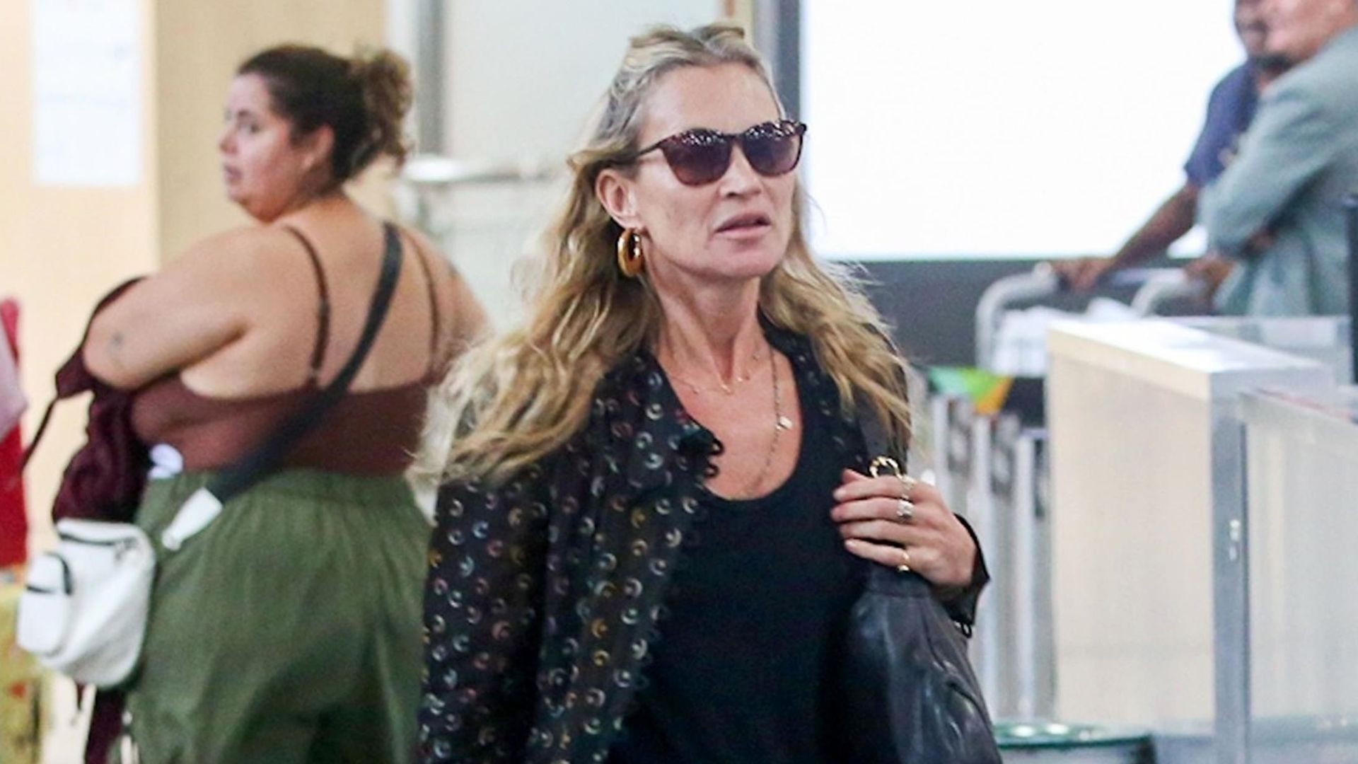 Kate Moss walks through the airport wearing black wide-leg jeans with a black vest top, a printed shirt and heeled boots.