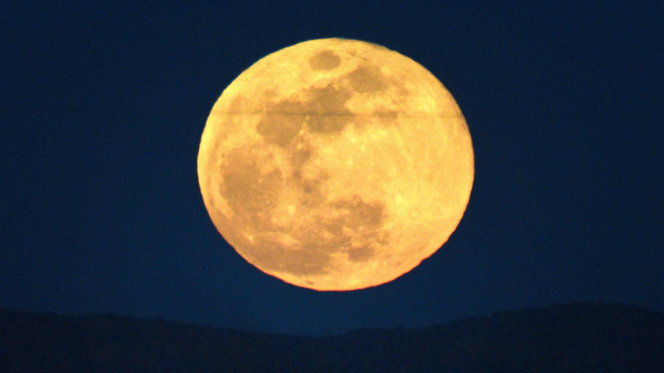 A yellow full moon is pictured rising in a dark sky above a silhouetted mountain ridge.