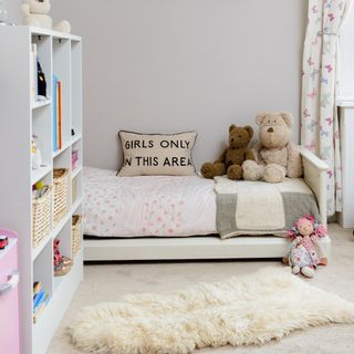 A childrens bedroom with a white bookcase and low profile white bed with a cushion that reads 'girls only in this area'.