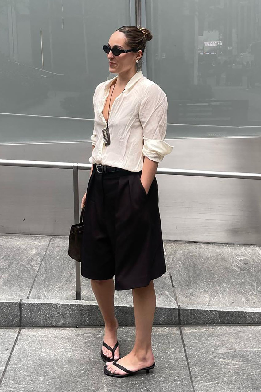 A woman wearing a white button-down shirt, black Bermuda shorts, and black kitten heel thong sandals.