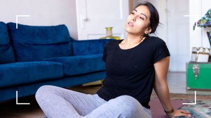 Woman sitting on yoga mat in living room next to sofa, wearing activewear with glass of water next to her, wondering whether you should exercise if you feel run down
