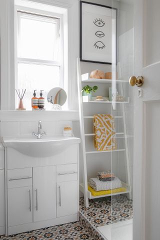 Small white bathroom with patterned tile flooring, vanity unit with cupboard doors, and ladder shelf housing towels and accessories