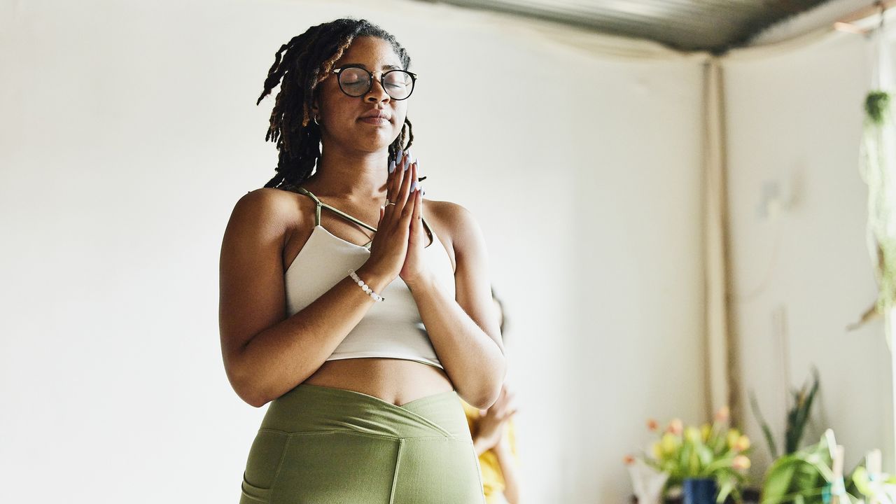 A young woman stands in a brightly lit room with her eyes closed and hands pressed together at her chest, performing the classic yoga prayer pose