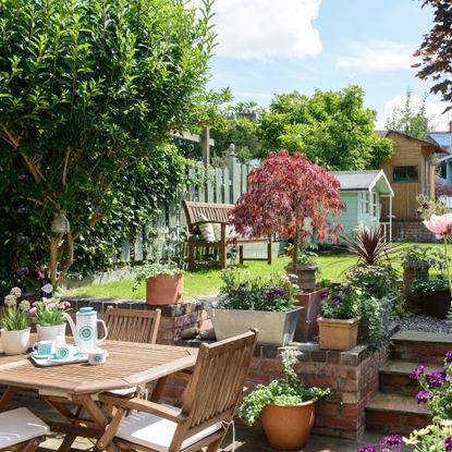 Garden in bloom with flowers, plants, steps and wooden table and chairs with blue shed in background