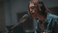 Harris Dickinson as John Lennon, singing into a microphone with long hair and glasses