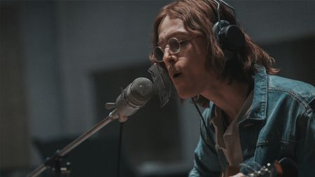 Harris Dickinson as John Lennon, singing into a microphone with long hair and glasses