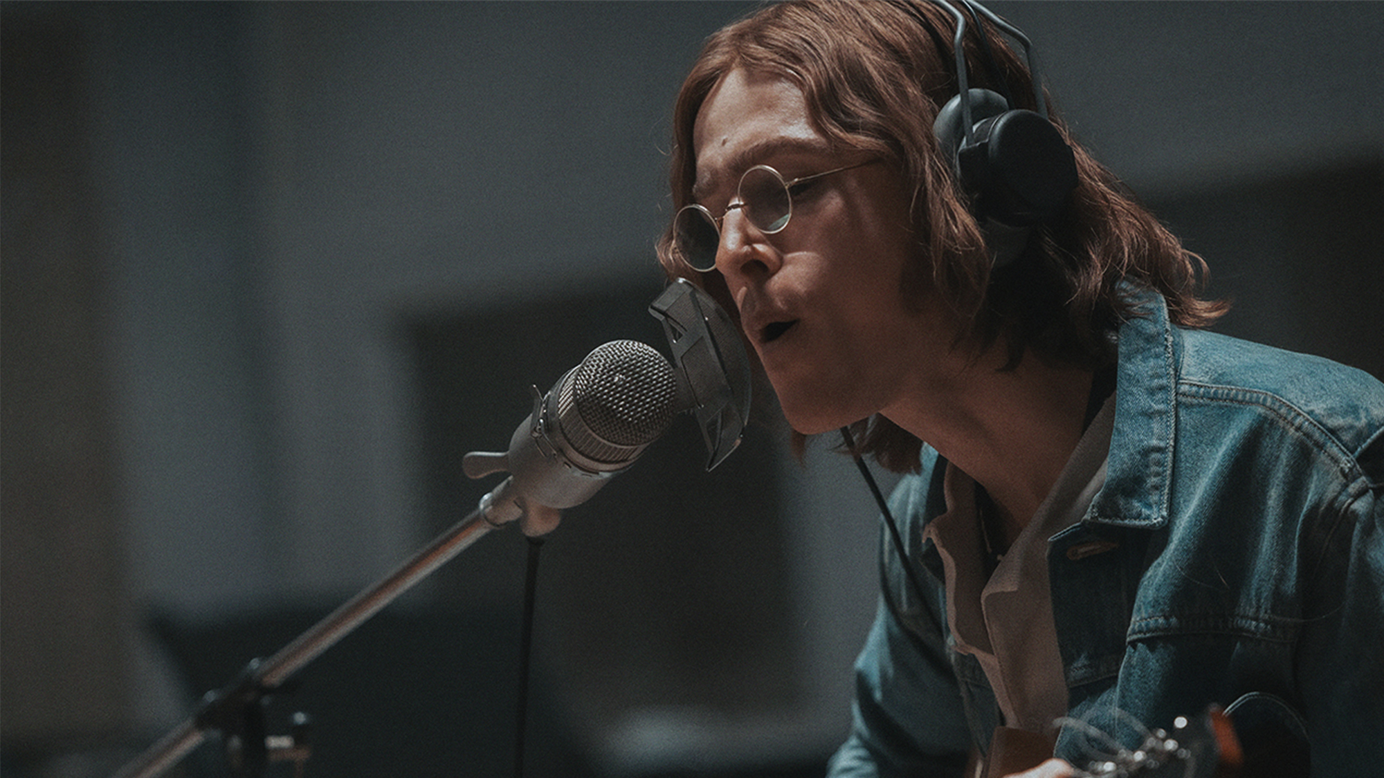 Harris Dickinson as John Lennon, singing into a microphone with long hair and glasses