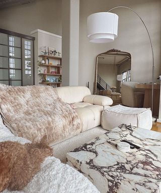 A neutral colored living room with a white couch with multiple neutral fluffy throws and marble table