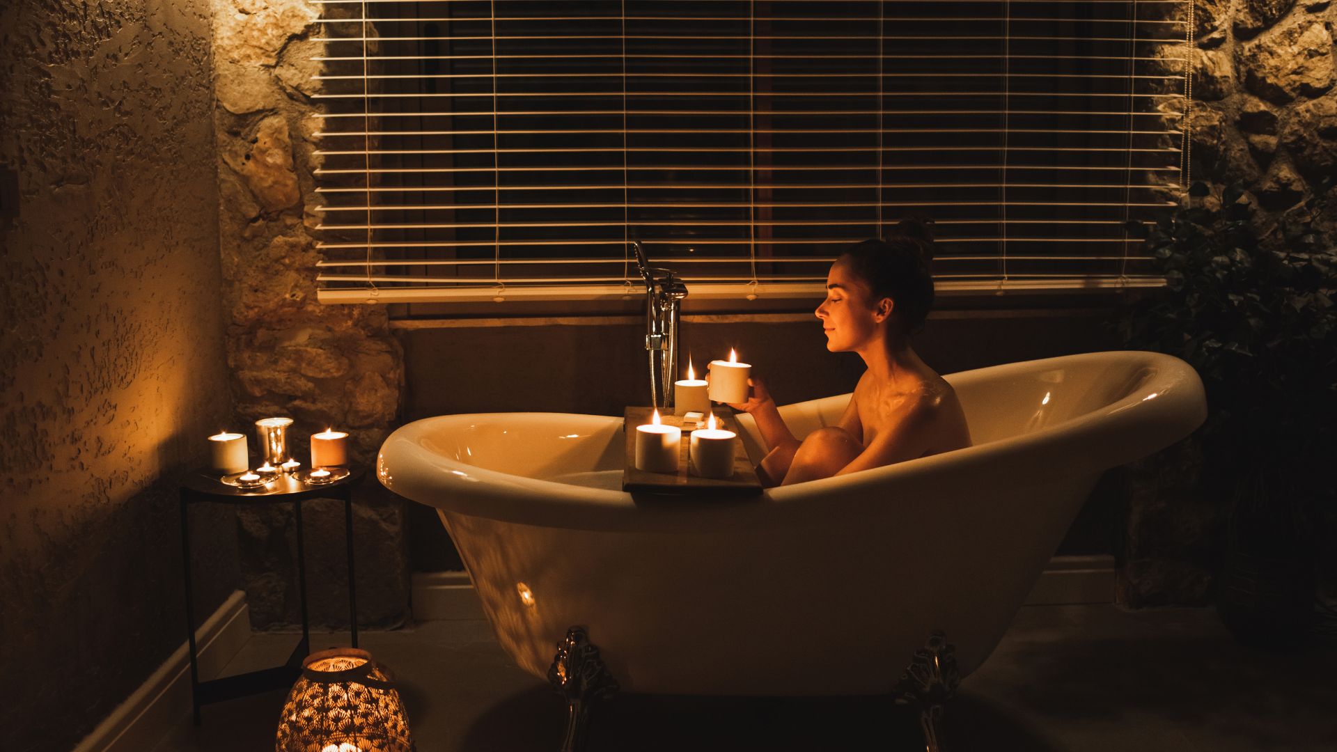 A woman sits up in a bath at night time. She is holding one of the candles that are in the room.