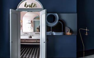 A room in the Henrietta Hotel. Deep blue walls, with a white door that leads to the bathroom. We see two sinks through the door and a big mirror. The walls are pink and the tiles on the floor are black and white.